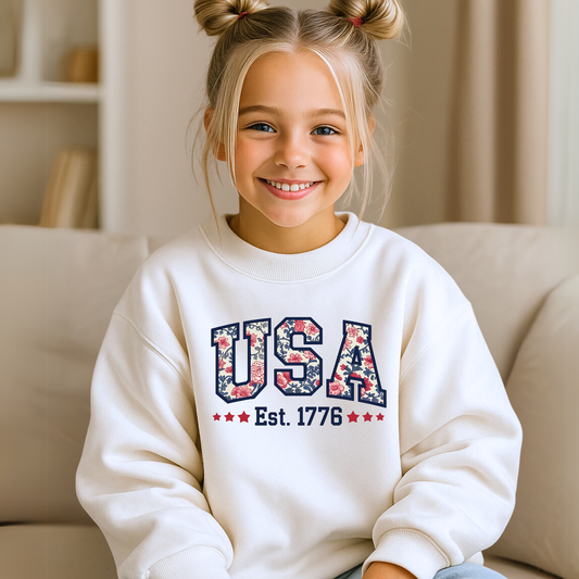 Girl in white USA Est. 1776 crewneck, seated and smiling on a beige couch.

