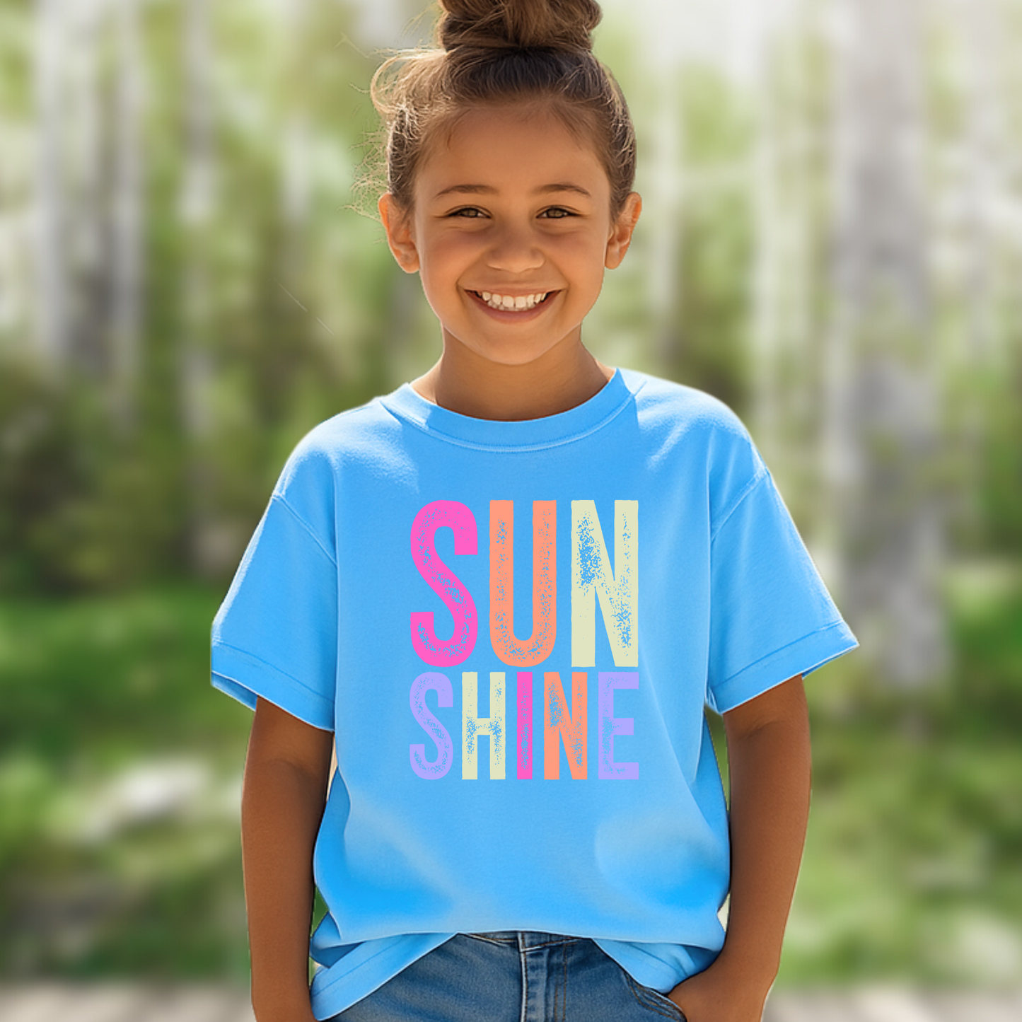 Girl outdoors in blue “Sunshine” tee with hands in pockets, smiling

