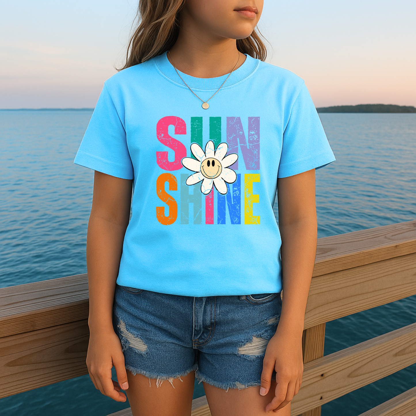 Girl standing on a dock wearing a light blue Sunshine shirt with a smiling daisy and colorful rainbow text

