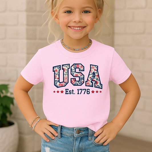 Girl wearing a light pink USA Est. 1776 youth t-shirt, smiling confidently in a casual outdoor  setting.

