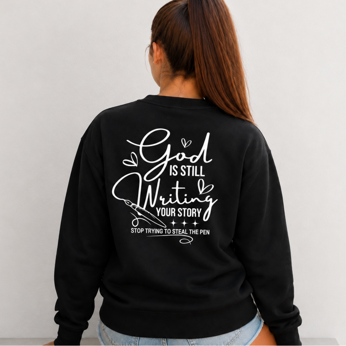 Back view of a woman wearing a black God Is Still Writing Your Story sweatshirt with white script lettering and a pen graphic.