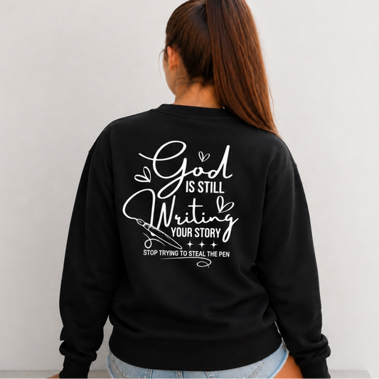 Back view of a woman wearing a black God Is Still Writing Your Story sweatshirt with white script lettering and a pen graphic.