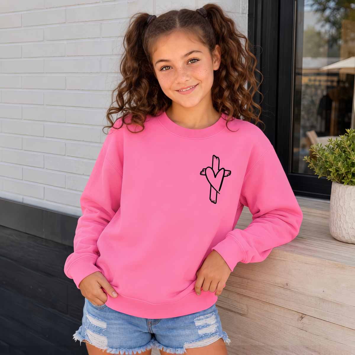 Girl wearing a Bright Pink God Is Still Writing Your Story youth crewneck sweatshirt with a small cross and heart graphic on the front chest.