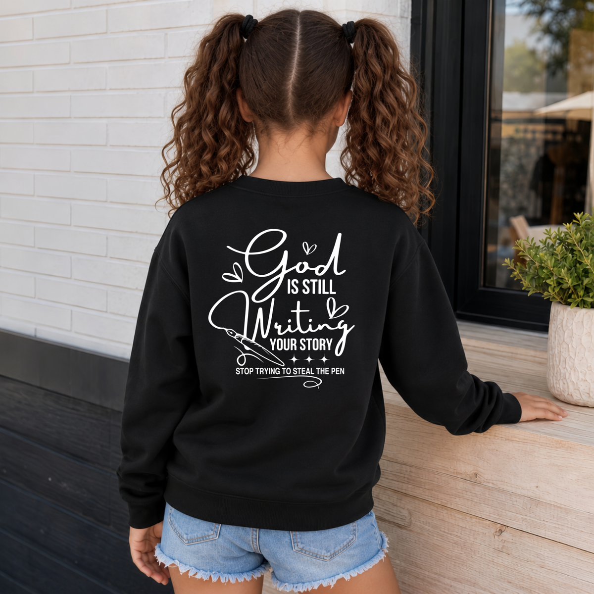 Back view of a girl wearing a black God Is Still Writing Your Story crewneck sweatshirt with white script lettering and a pen graphic.