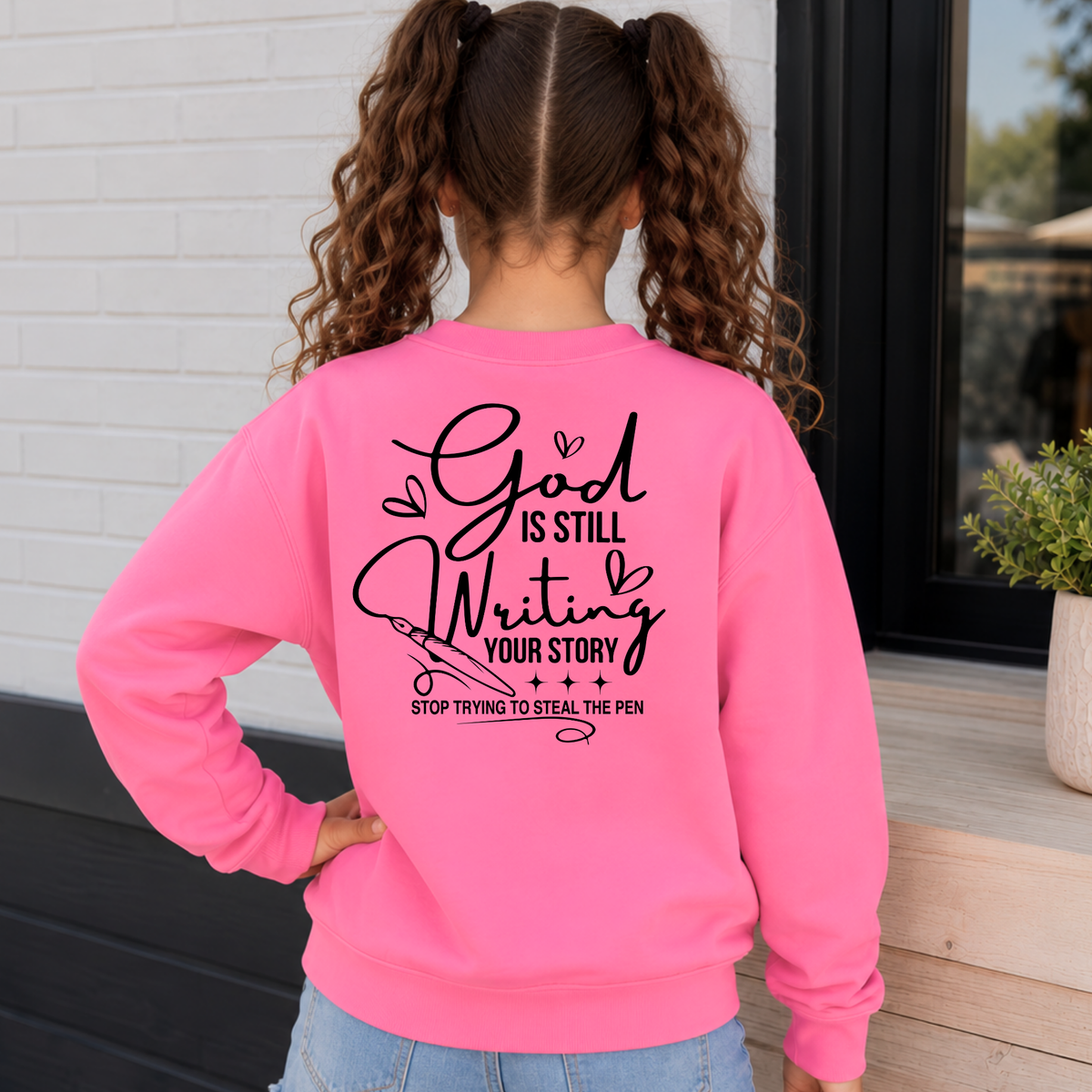 Back view of a girl wearing a Bright Pink God Is Still Writing Your Story crewneck sweatshirt with black script lettering and a pen graphic.
