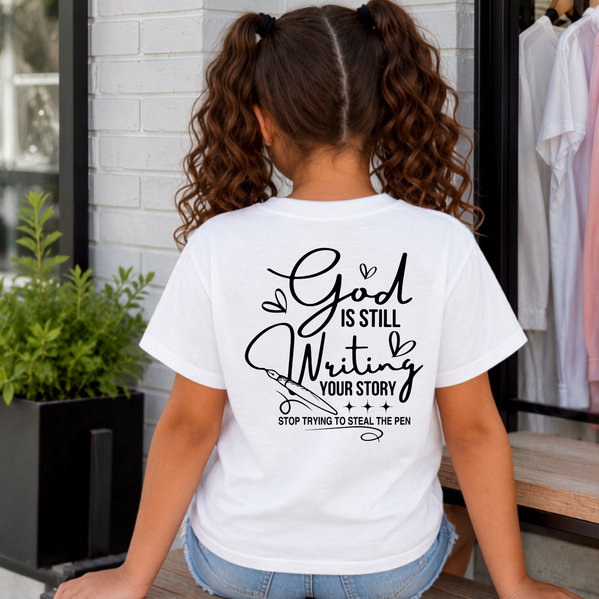 Back view of a girl wearing a white God Is Still Writing Your Story T-shirt with black script lettering and a pen graphic.