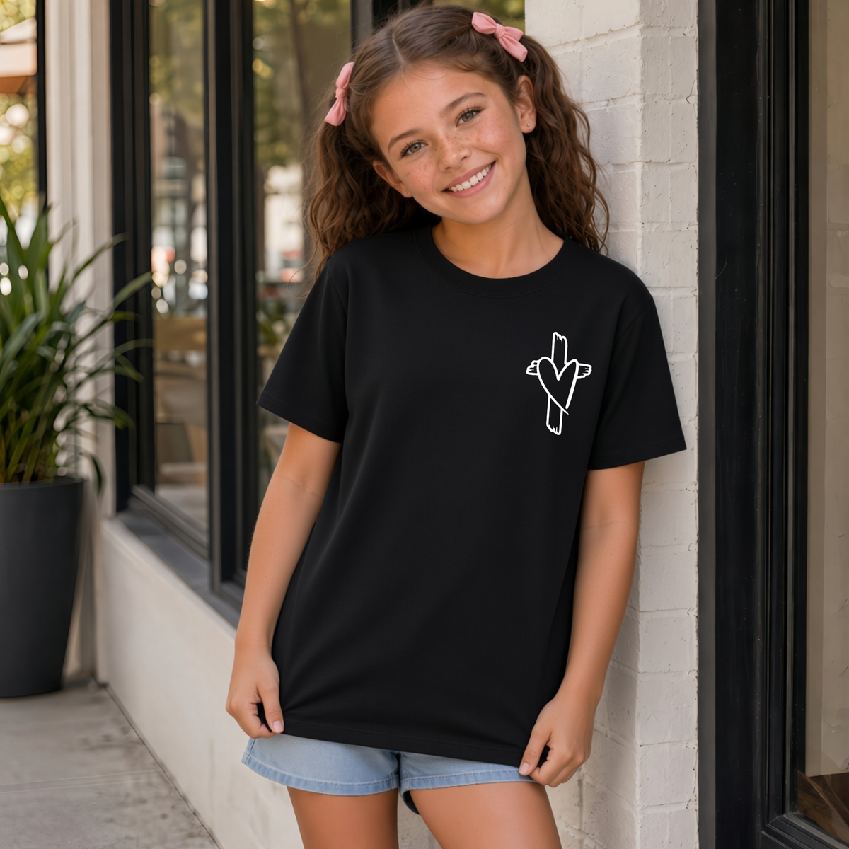 Girl wearing a black God Is Still Writing Your Story youth T-shirt with a small cross and heart graphic on the front chest.