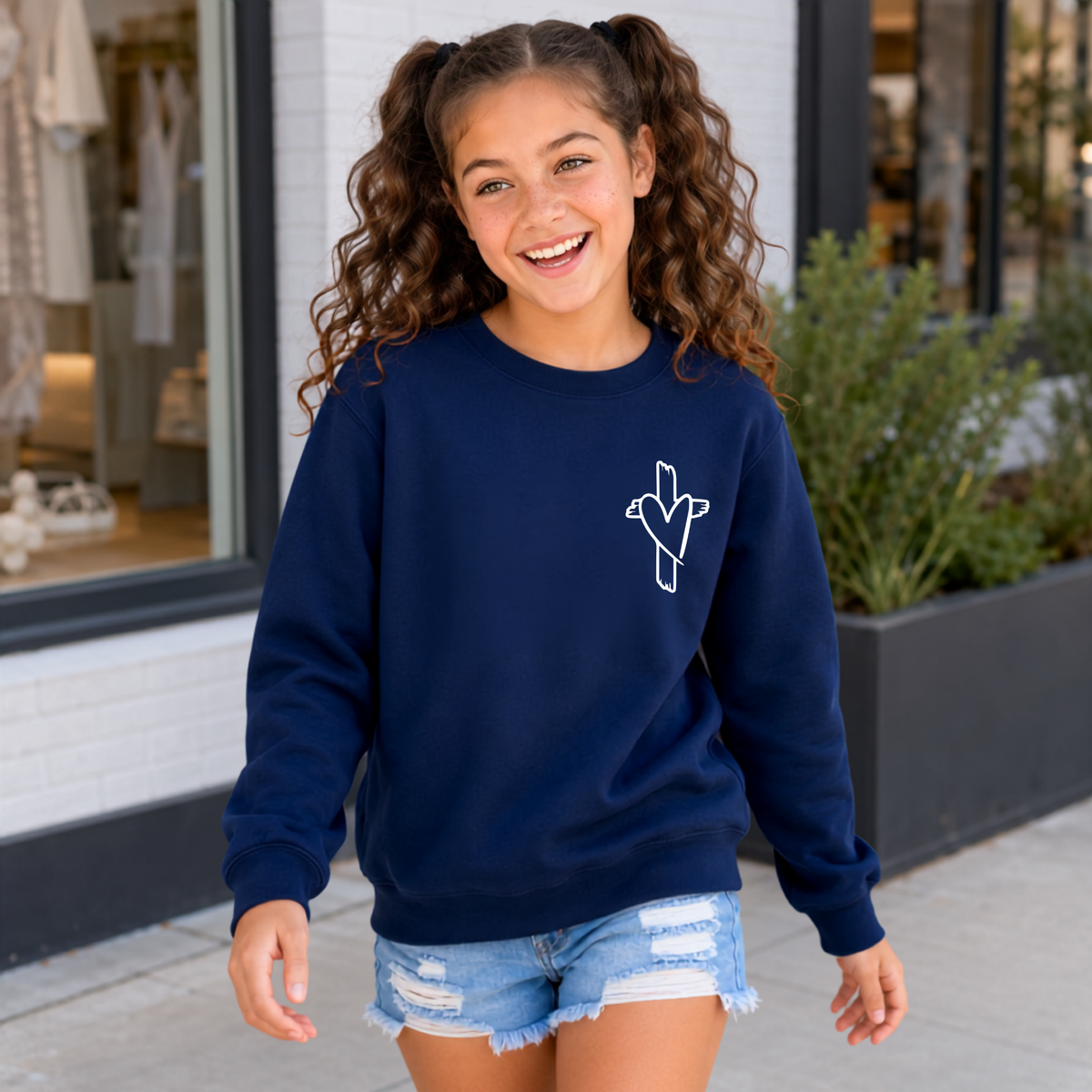 Girl wearing a navy God Is Still Writing Your Story crewneck sweatshirt with a small cross and heart graphic on the front chest.
