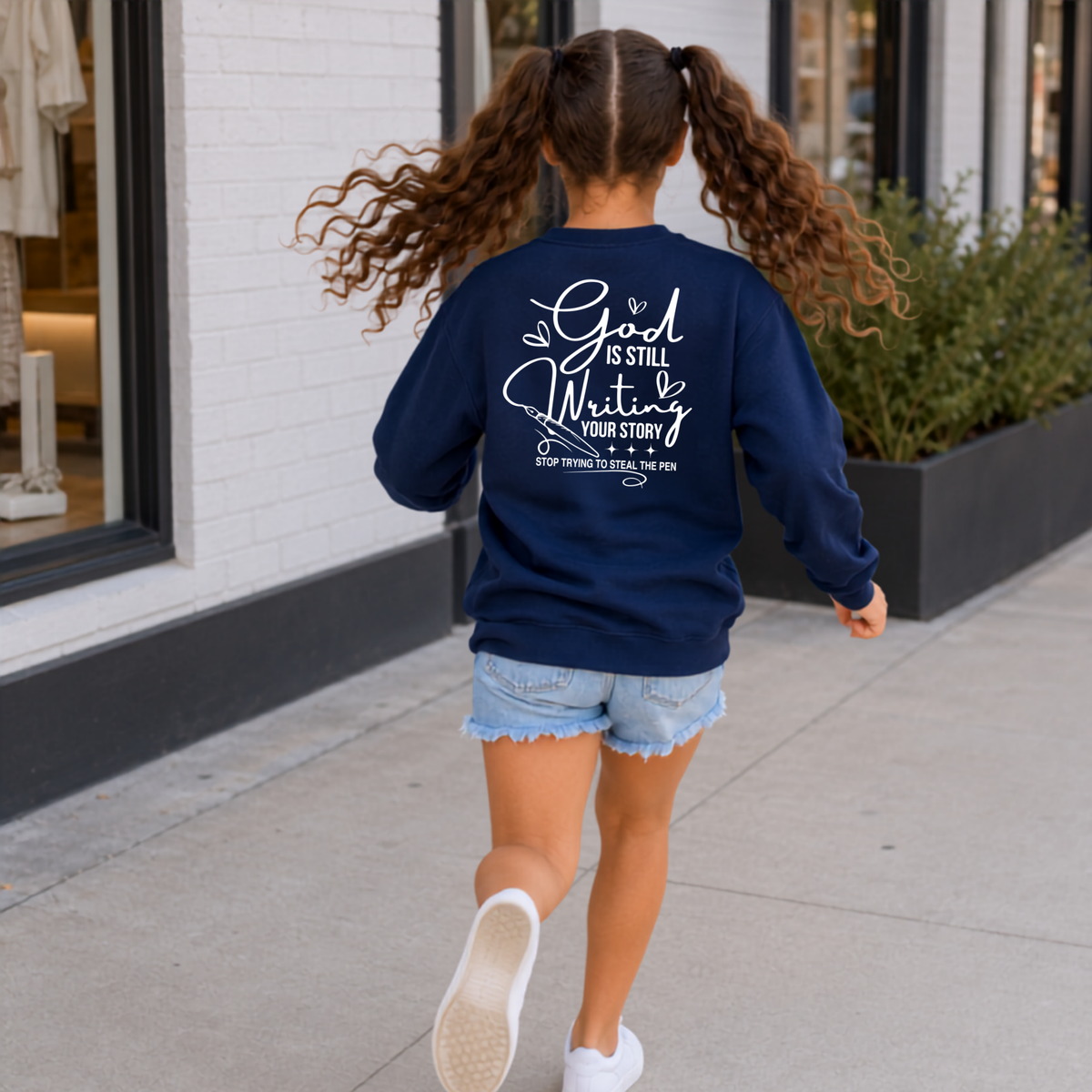Back view of a woman wearing a navy God Is Still Writing Your Story crewneck sweatshirt with white script lettering and a pen graphic.