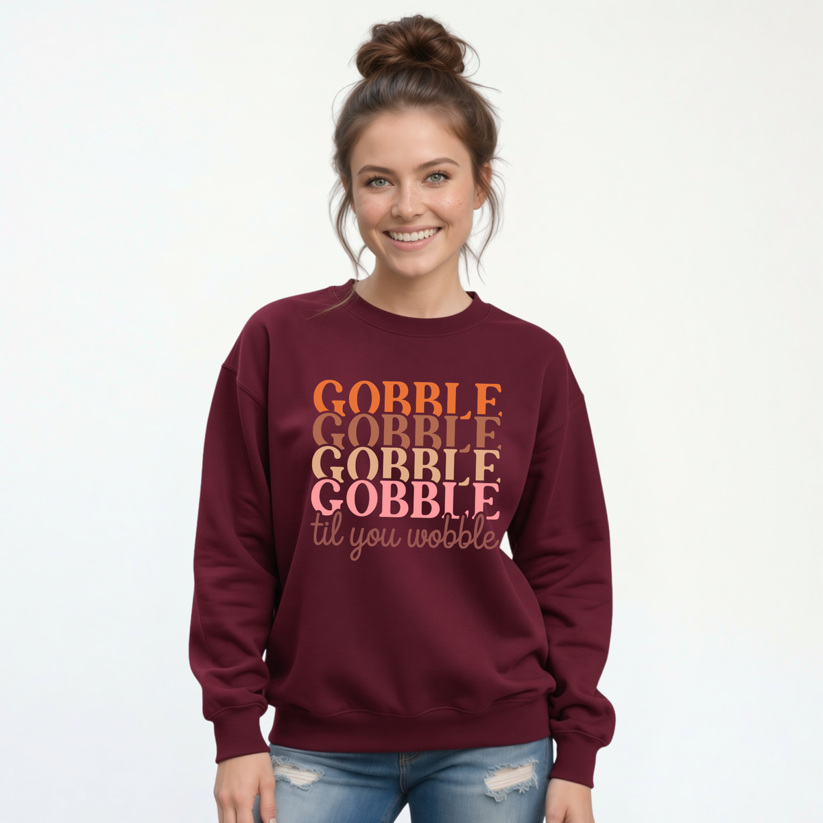 Woman in a maroon crewneck sweatshirt featuring the “Gobble Gobble Gobble Gobble til you wobble” Thanksgiving design with distressed jeans.
