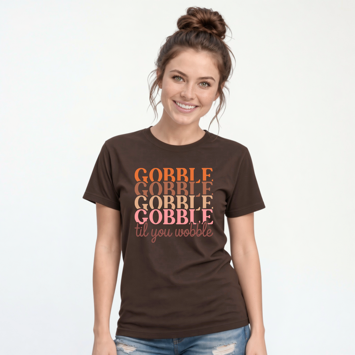 Woman modeling a brown T-shirt with the colorful stacked “Gobble Gobble Gobble Gobble til you wobble” Thanksgiving design and blue jeans.