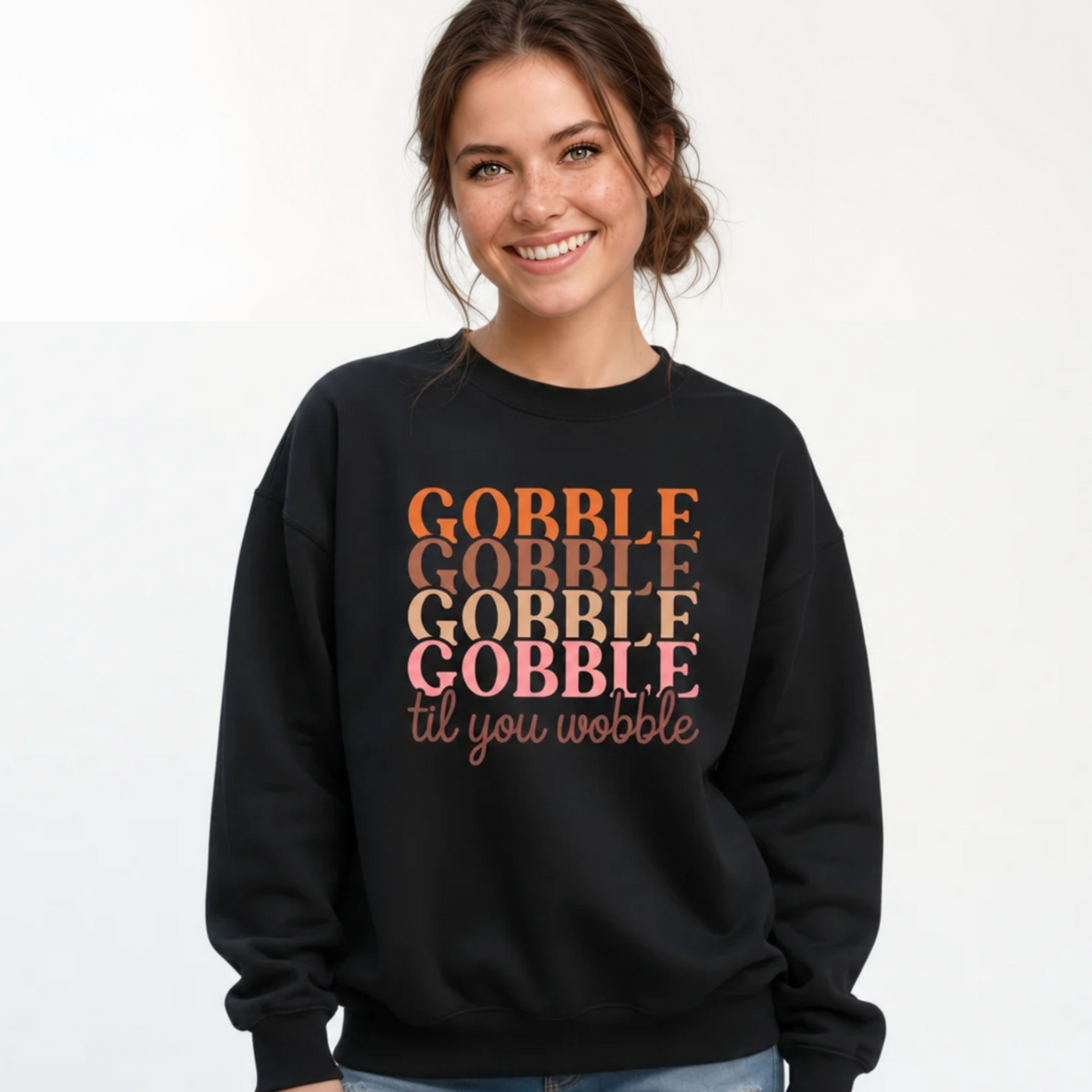A woman smiling and wearing a black crewneck sweatshirt with the text 'GOBBLE GOBBLE GOBBLE til you wobble' printed on the front in orange, brown, and pink tones, paired with blue jeans against a plain white background.