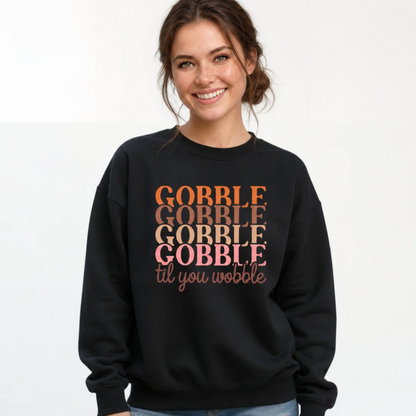 A woman smiling and wearing a black crewneck sweatshirt with the text 'GOBBLE GOBBLE GOBBLE til you wobble' printed on the front in orange, brown, and pink tones, paired with blue jeans against a plain white background.