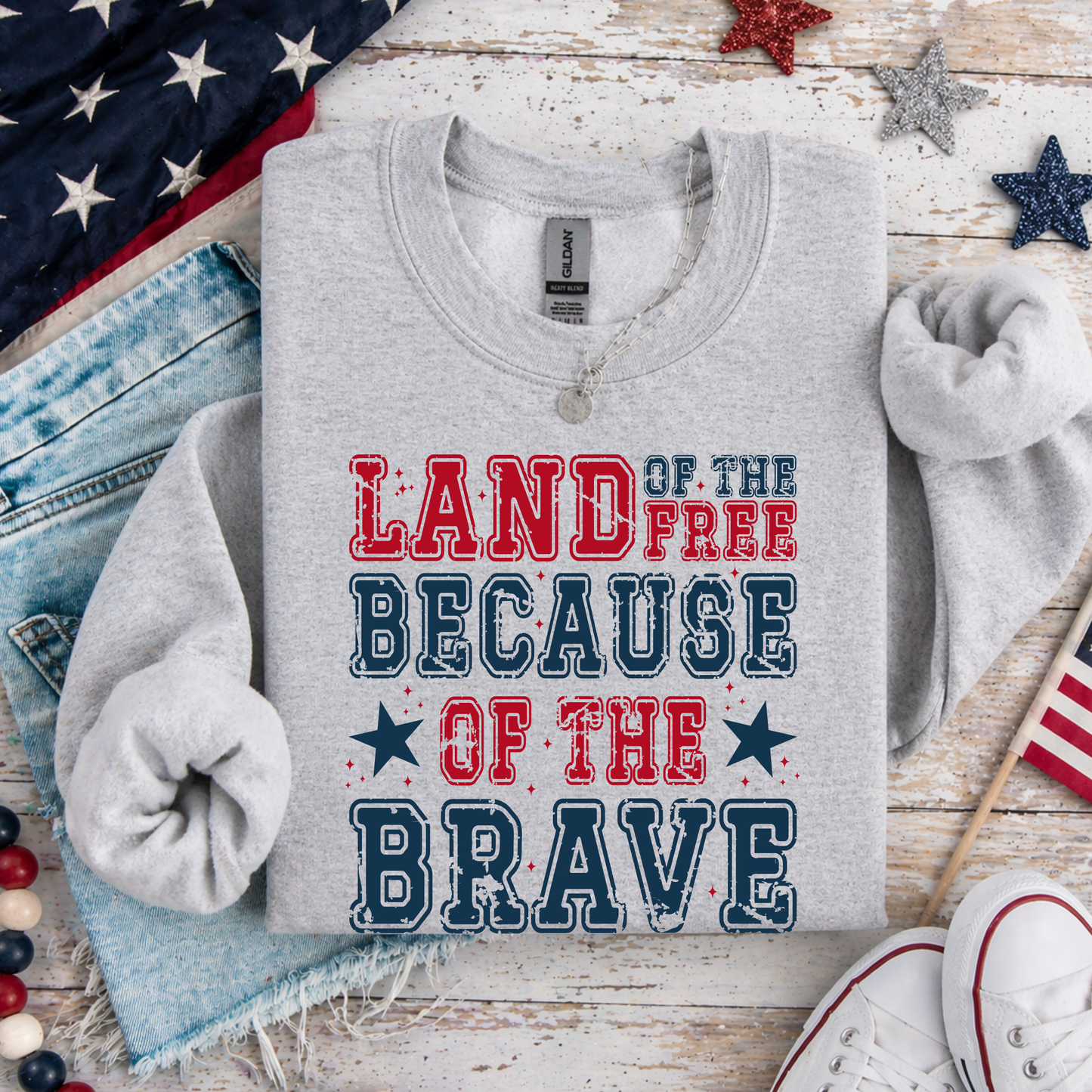 Gray sweatshirt with 'Land of the Free Because of the Brave' text on a wooden surface with American flags and stars.