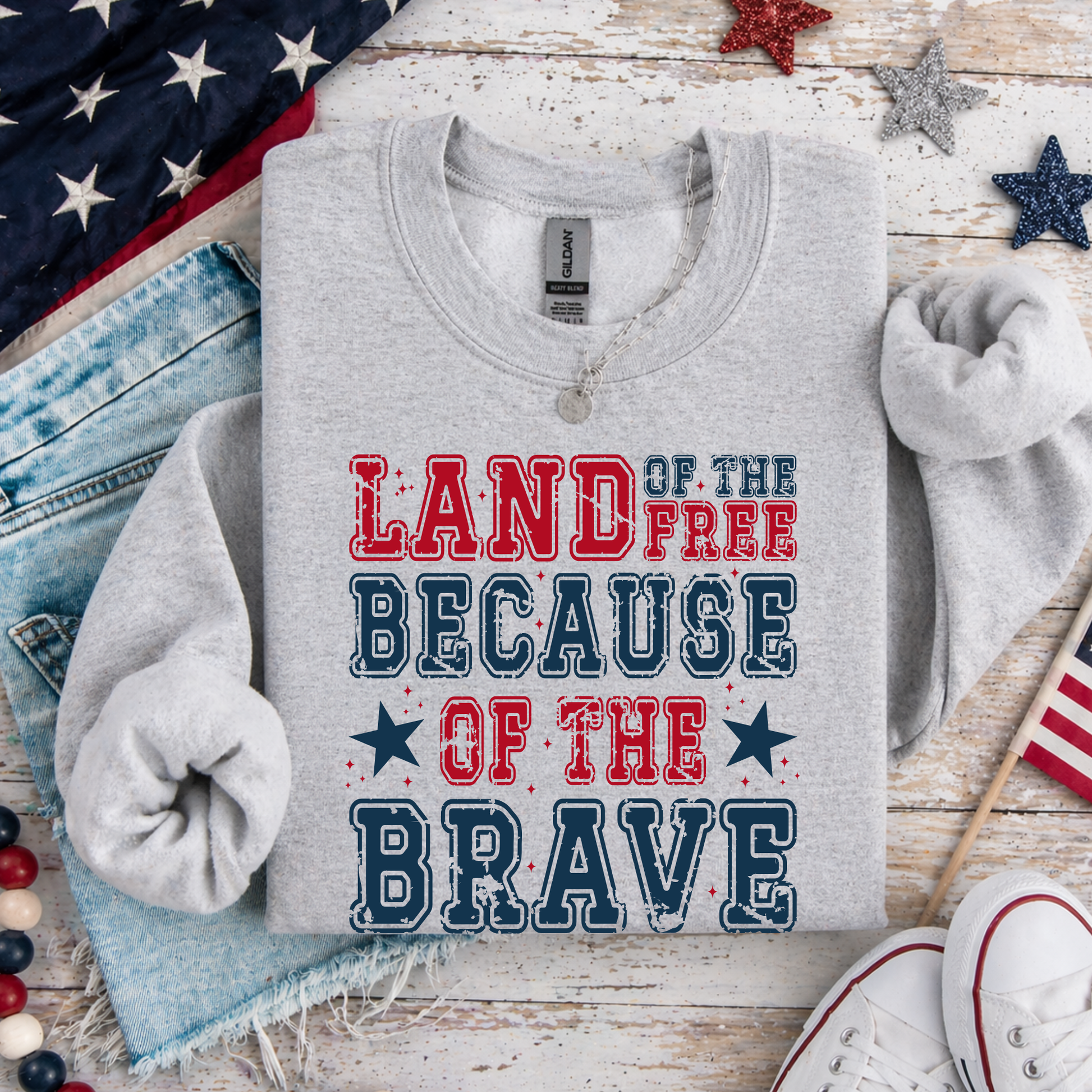 Gray sweatshirt with 'Land of the Free Because of the Brave' text on a wooden surface with American flags and stars.