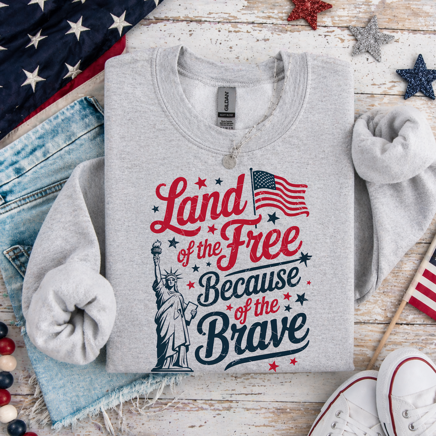 Gray sweatshirt with 'Land of the Free Because of the Brave' text and Statue of Liberty graphic on a wooden surface with American flags and stars.