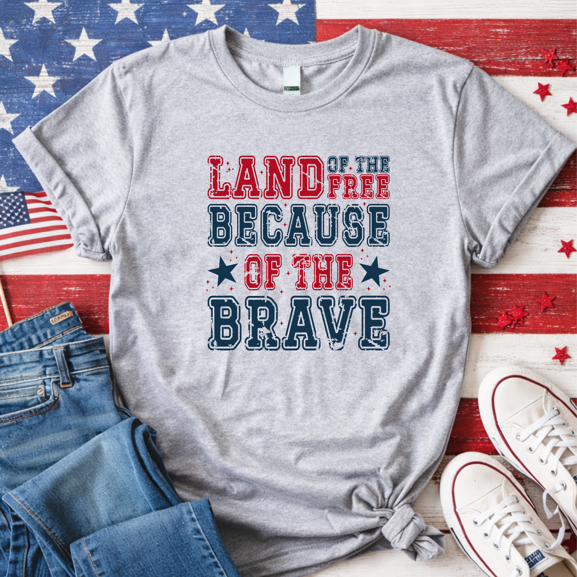 Gray t-shirt with 'Land of the Free Because of the Brave' text on a wooden surface with American flags and shoes.