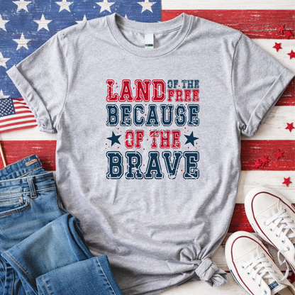 Gray t-shirt with 'Land of the Free Because of the Brave' text on a wooden surface with American flags and shoes.