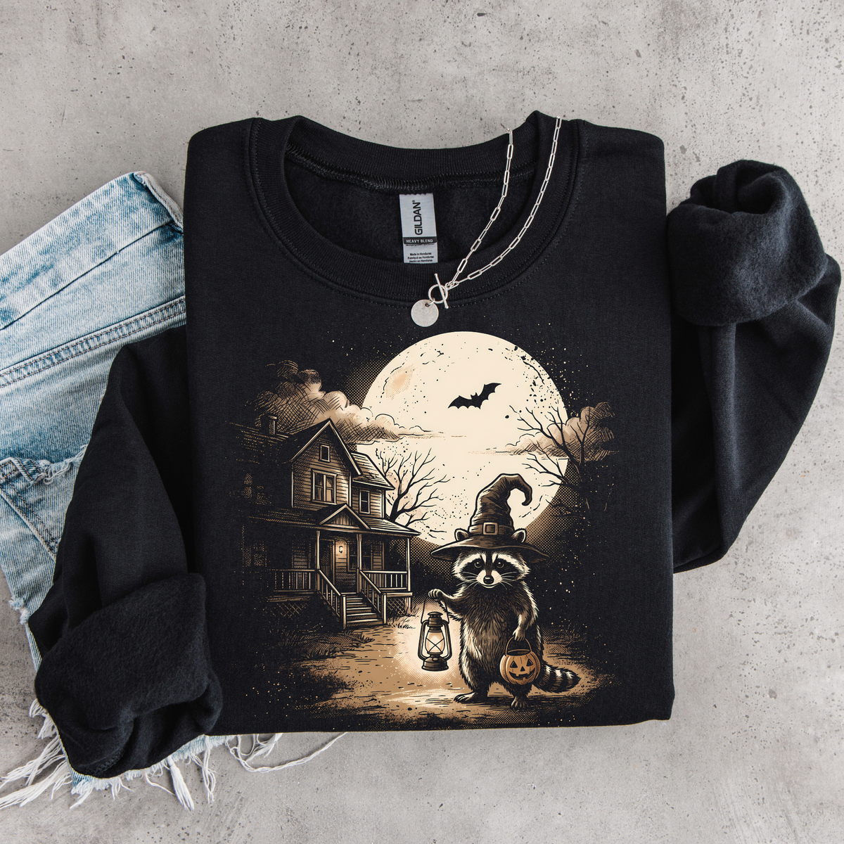 Black sweatshirt flat lay featuring a raccoon witch, jack-o'-lantern, bats and full moon Halloween scene.