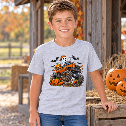 Boy wearing a sports gray youth T-shirt with an orange Halloween monster truck, friendly ghost, jack-o-lanterns, and bats beside hay bales and carved pumpkins.