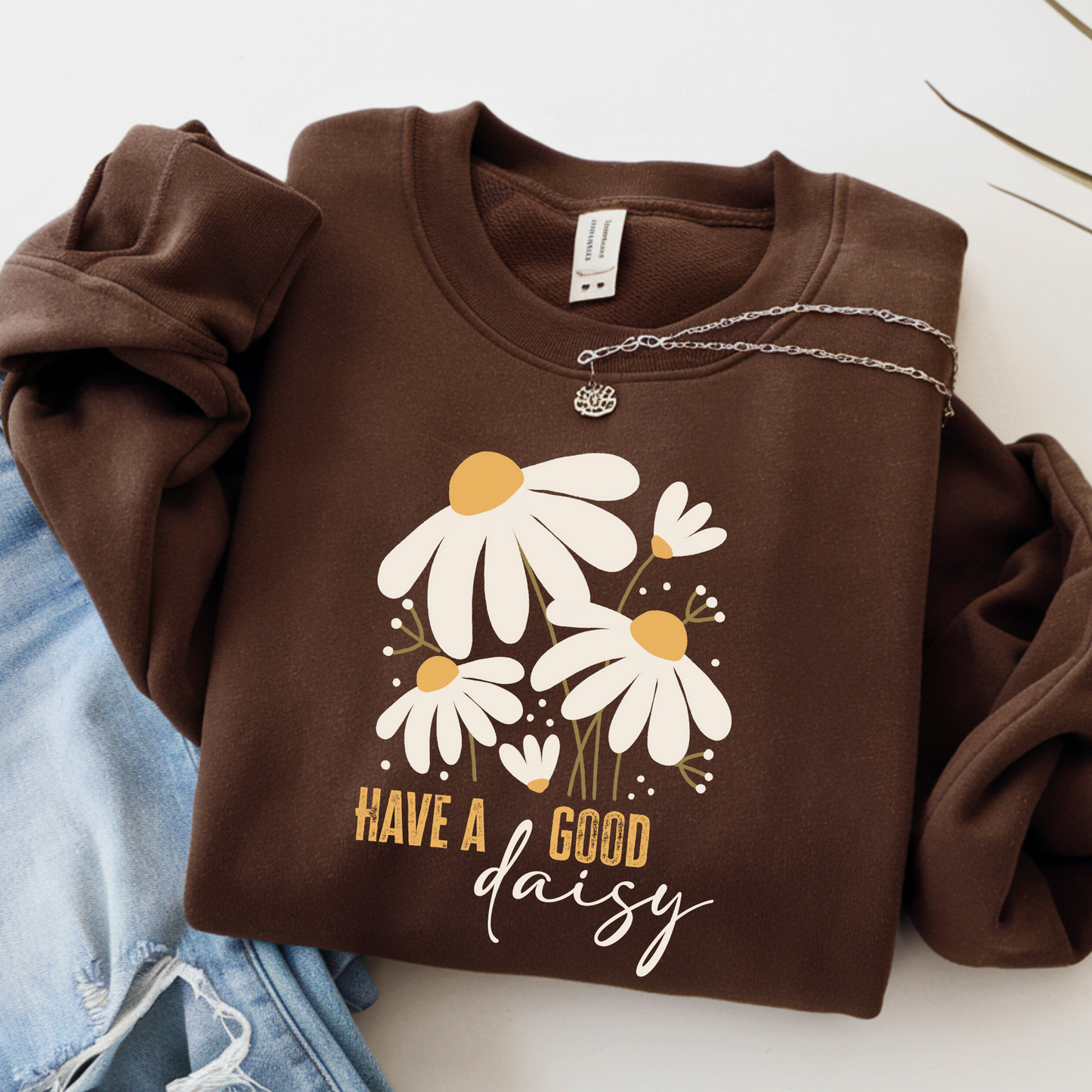 Chocolate brown “Have a Good Daisy” sweatshirt folded with jeans and a necklace, featuring a cozy fall floral graphic perfect for seasonal outfits, pumpkin patch days, or casual autumn layering.

