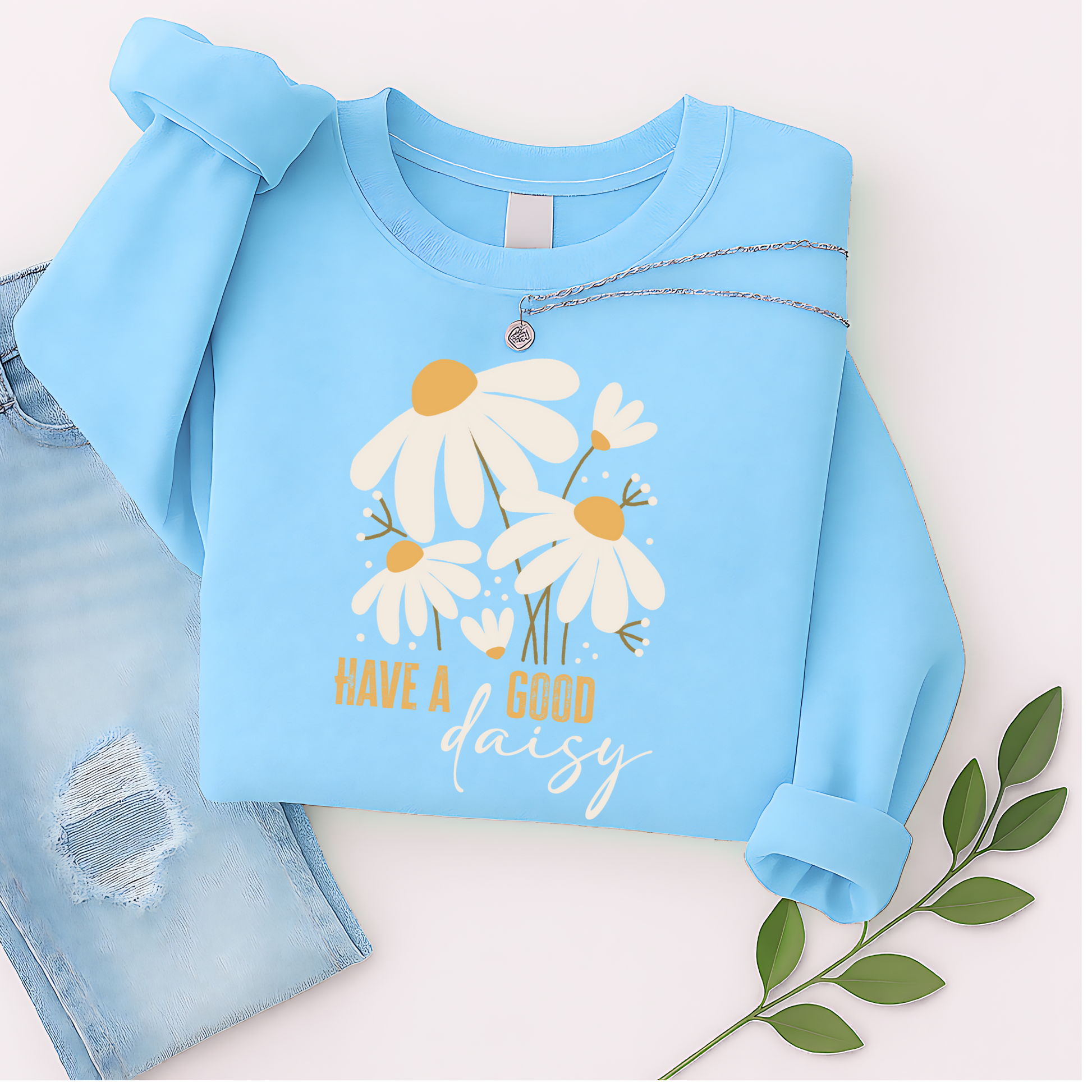 Sky blue floral sweatshirt with “Have a Good Daisy” design, laid out with jeans and greenery for a light, playful fall flat lay—perfect for transitional weather and cheerful cozy vibes.