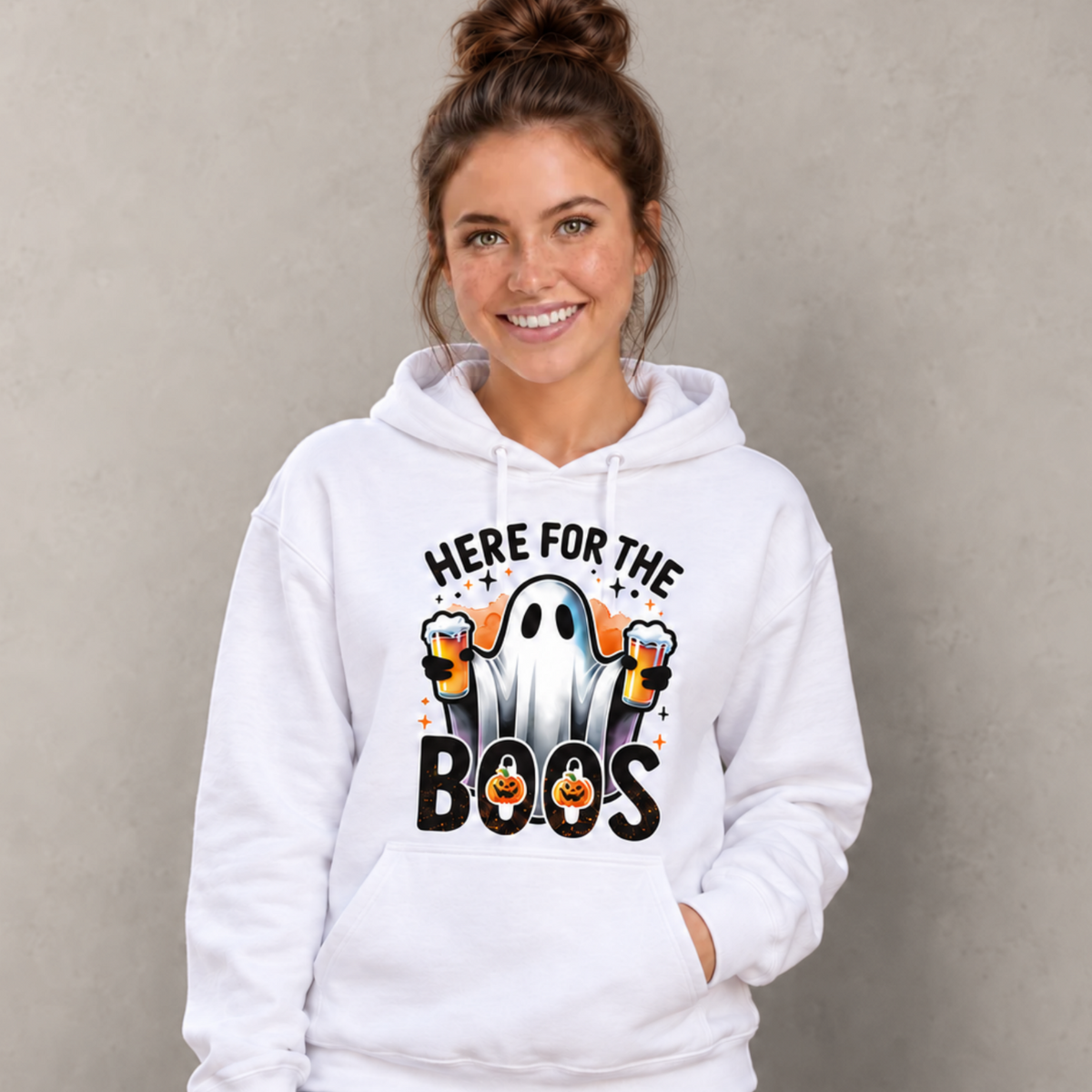 Woman wearing a white hoodie with a ghost holding two beer mugs and Here For The Boos Halloween design on a gray background.