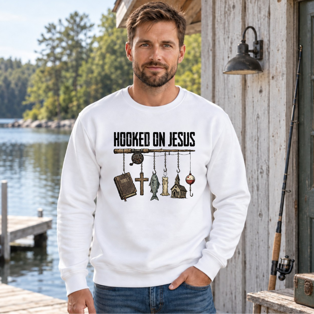 Man wearing a white Hooked on Jesus sweatshirt with fishing rod, Bible, cross, fish, candle, church, and bobber graphic outside a lakeside cabin.