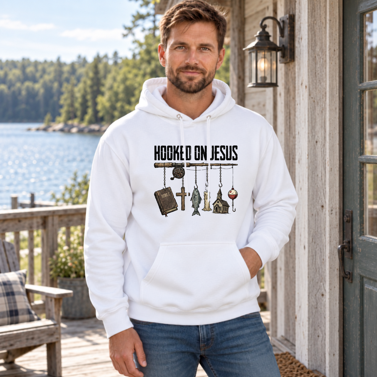 Man wearing a white Hooked on Jesus hoodie beside a rustic lakeside cabin.