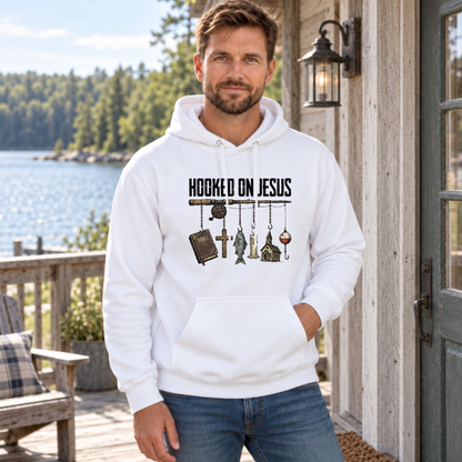 Man wearing a white Hooked on Jesus hoodie beside a rustic lakeside cabin.
