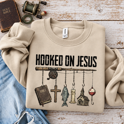 Folded sand crewneck sweatshirt with Hooked on Jesus fishing design on whitewashed wood beside a Bible and fishing reel