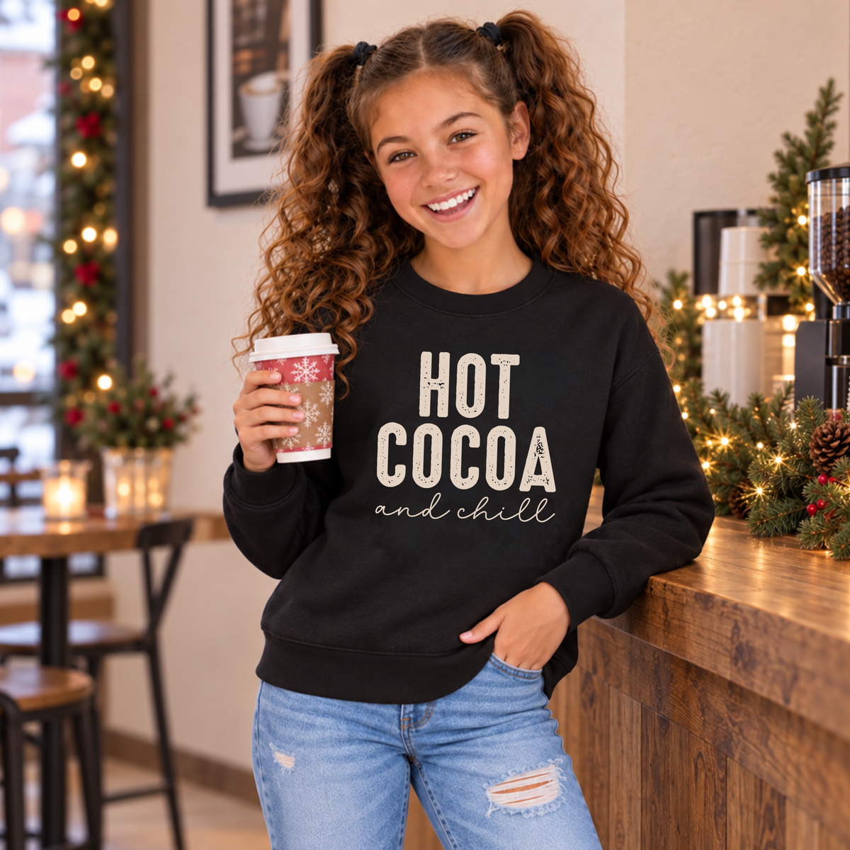 Youth girl wearing a black Hot Cocoa and Chill sweatshirt while holding a holiday cup in a festive cafe