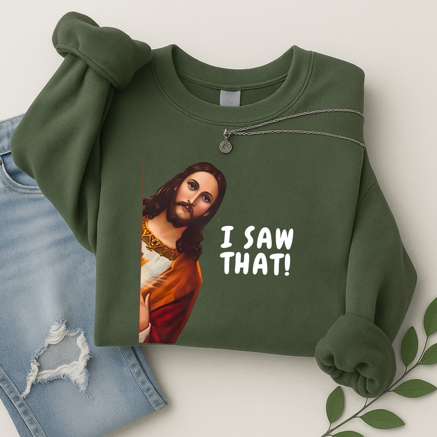 Green sweatshirt with a graphic of a Jesus saying 'I SAW THAT!' text, paired with jeans and leaves on a light background.