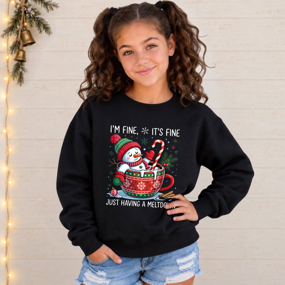 Girl wearing a black youth sweatshirt with an I'm Fine It's Fine snowman meltdown Christmas design against a light wood wall