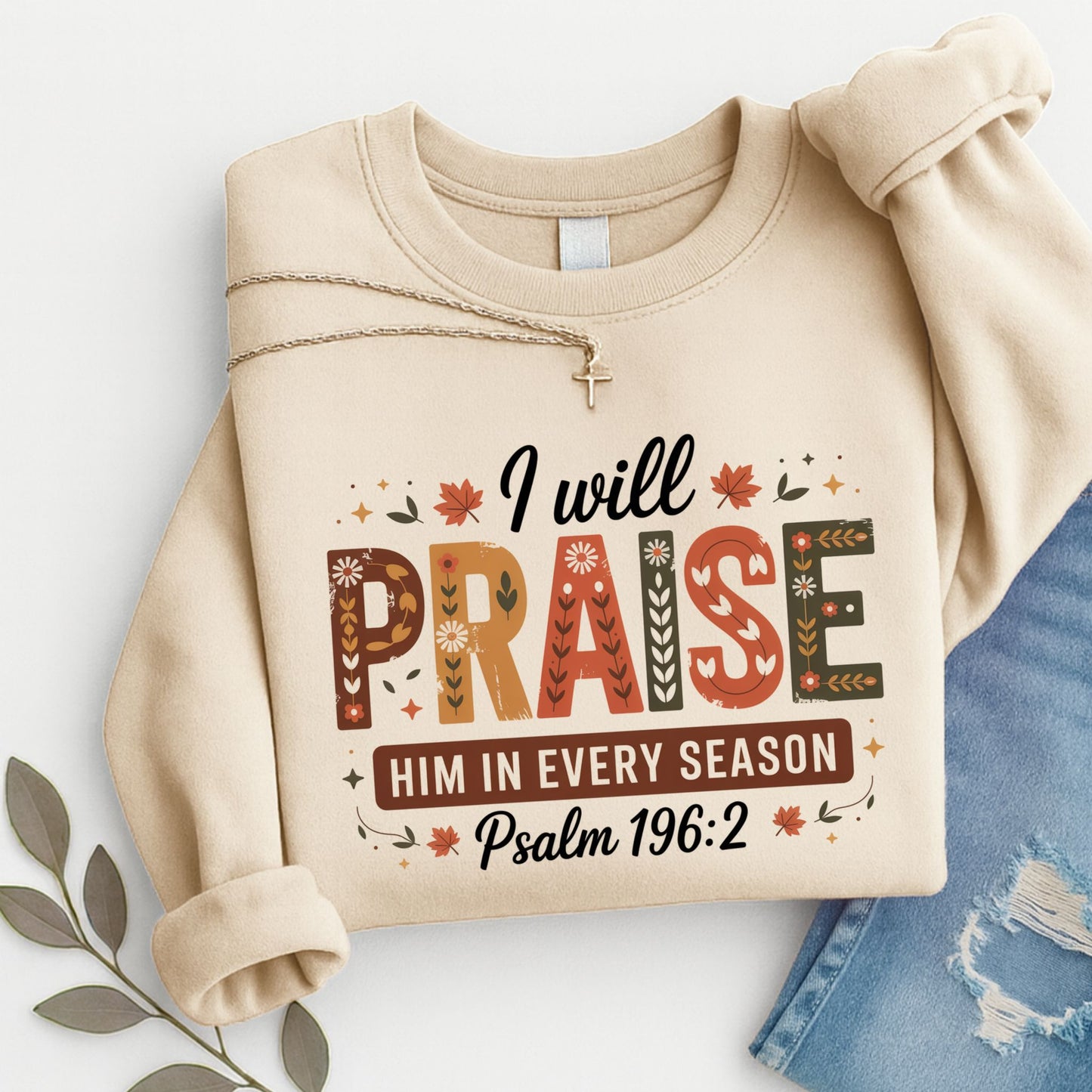 Folded sand “I Will Praise Him In Every Season” sweatshirt with cross necklace and jeans flat lay