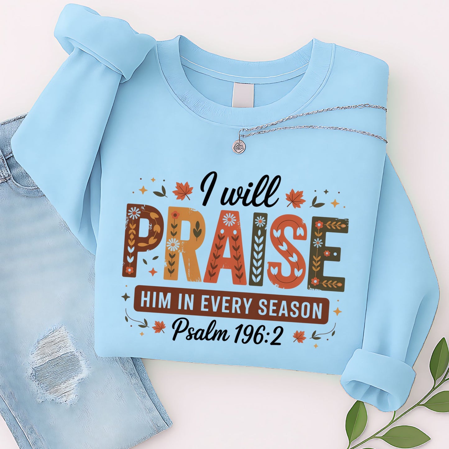 Folded light blue “I Will Praise Him In Every Season” sweatshirt styled with necklace and jeans flat lay
