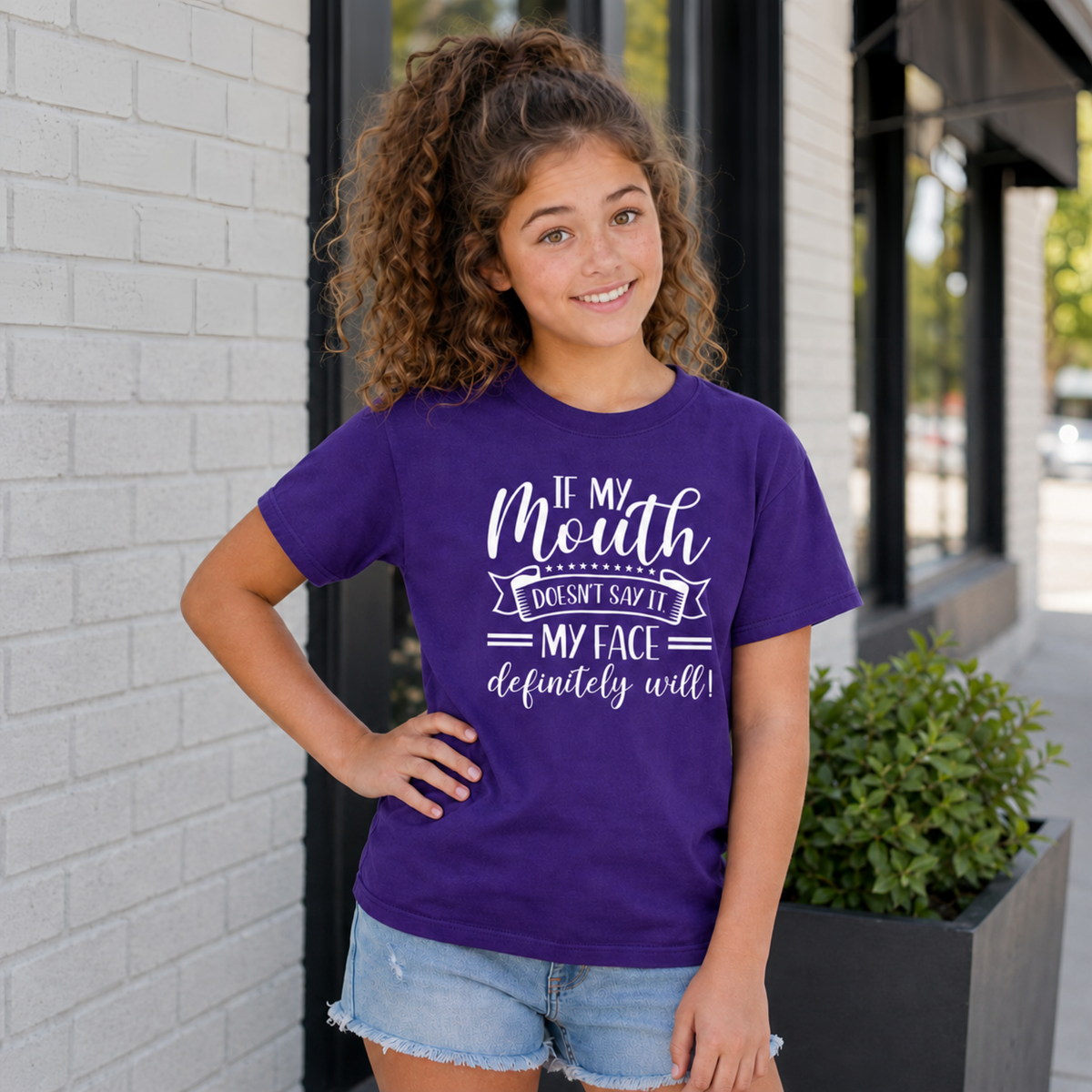 Youth model wearing a purple T-shirt with white “If My Mouth Doesn’t Say It, My Face Definitely Will” lettering outside a storefront.