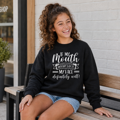 Laughing youth model wearing a black crewneck sweatshirt with white “If My Mouth Doesn’t Say It, My Face Definitely Will” lettering outside a storefront.