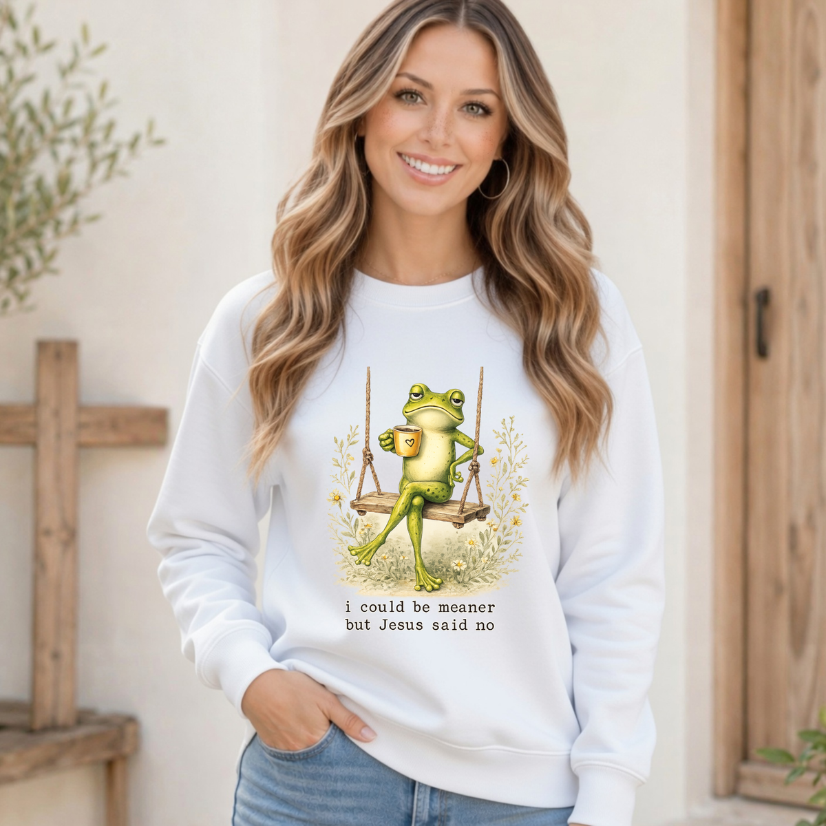 Woman modeling a white crewneck sweatshirt with a frog sitting on a swing holding a mug and the words “I could be meaner but Jesus said no.”