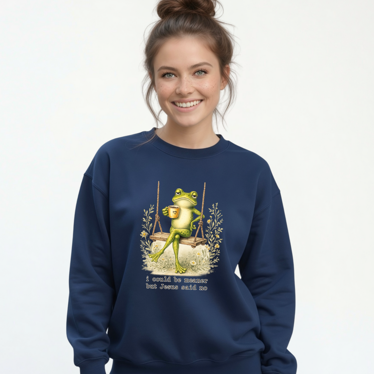 Woman modeling a navy crewneck sweatshirt with a frog sitting on a swing holding a mug and the words “I could be meaner but Jesus said no.”