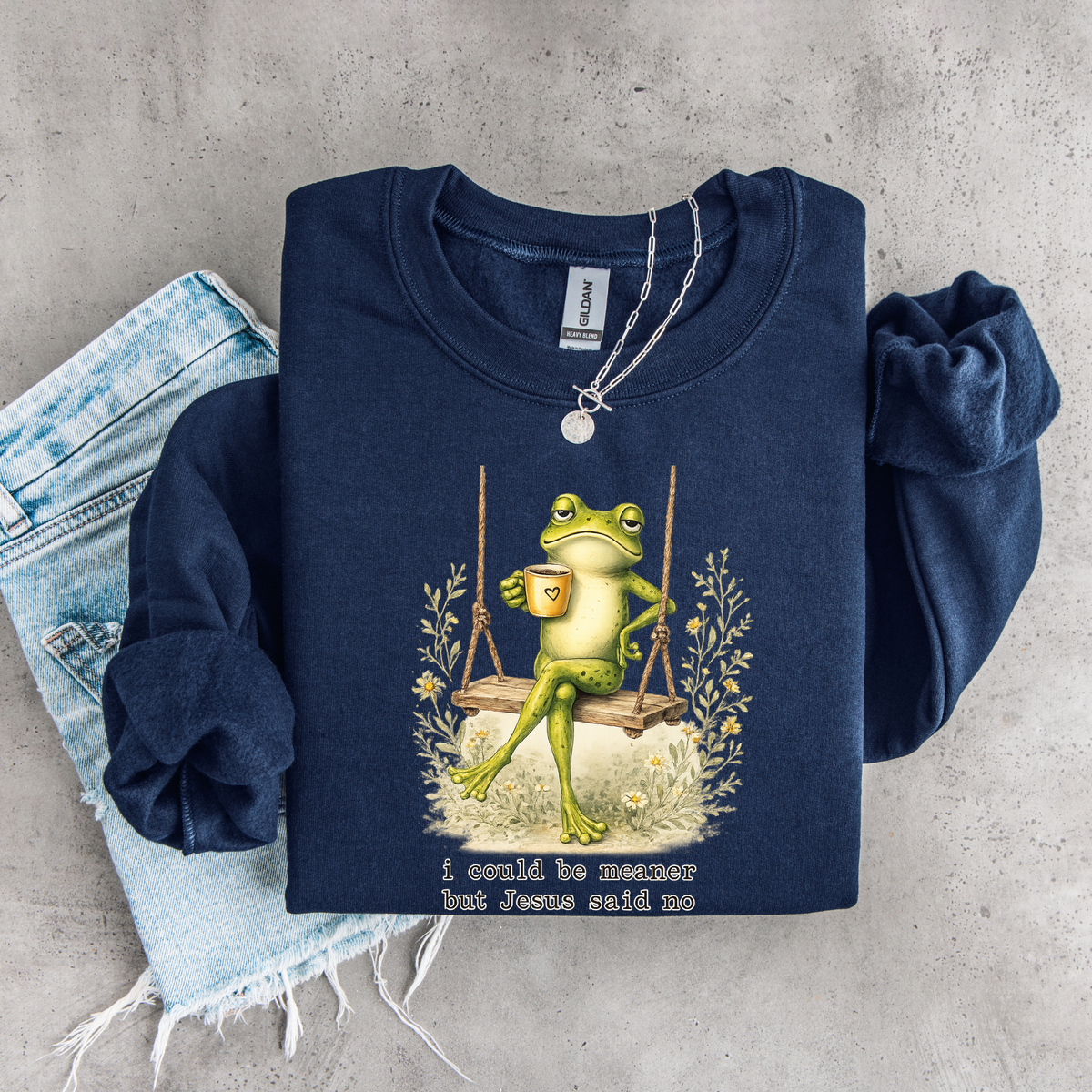 Folded navy crewneck sweatshirt flat lay with the I Could Be Meaner But Jesus Said No frog-on-a-swing faith design.
