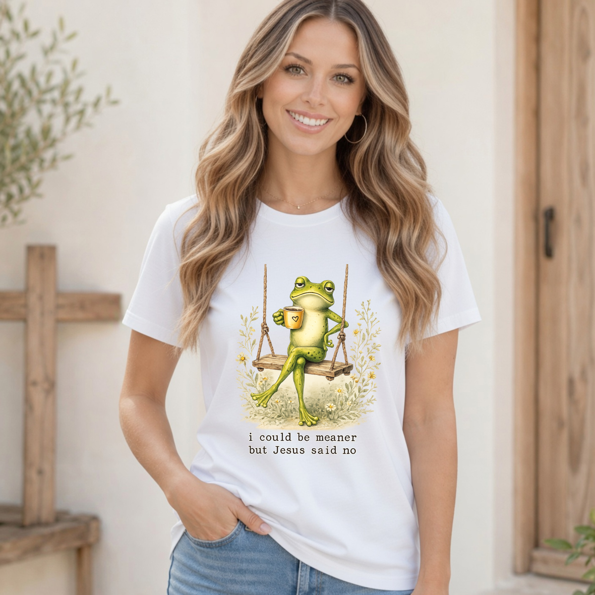 Woman modeling a white T-shirt with a frog sitting on a swing holding a mug and the words “I could be meaner but Jesus said no.”