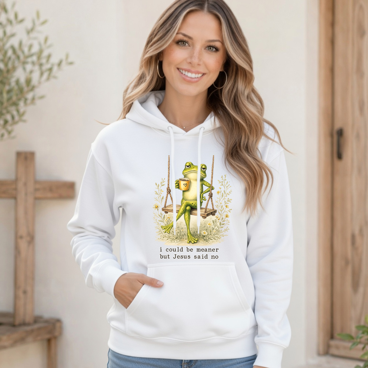 Woman modeling a white hoodie with a frog sitting on a swing holding a mug and the words “I could be meaner but Jesus said no.”