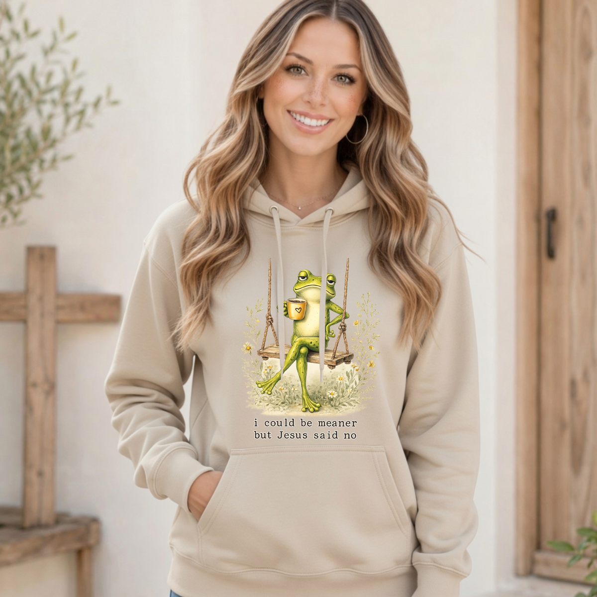 Woman modeling a sand hoodie with a frog sitting on a swing holding a mug and the words “I could be meaner but Jesus said no.”