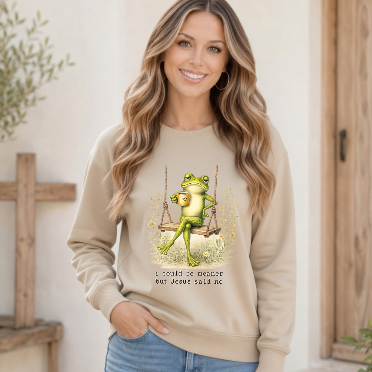 Woman modeling a sand crewneck sweatshirt with a frog sitting on a swing holding a mug and the words “I could be meaner but Jesus said no.”