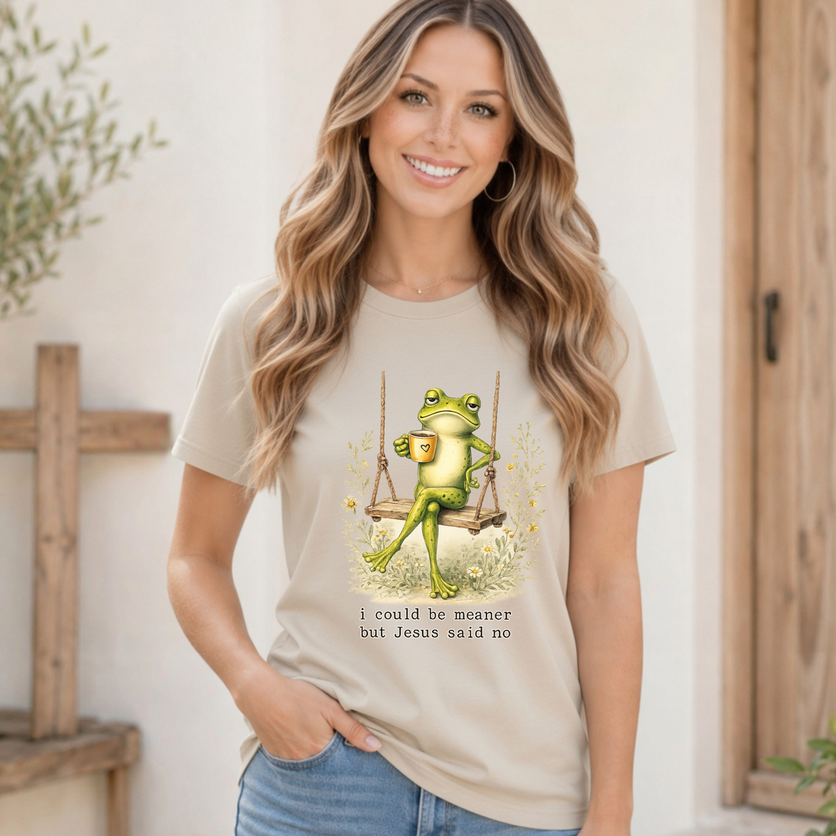 Woman modeling a sand T-shirt with a frog sitting on a swing holding a mug and the words “I could be meaner but Jesus said no.”