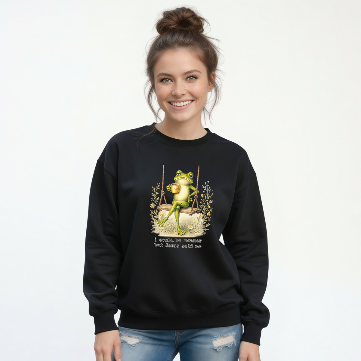 Woman modeling a black crewneck sweatshirt with a frog sitting on a swing holding a mug and the words “I could be meaner but Jesus said no.”
