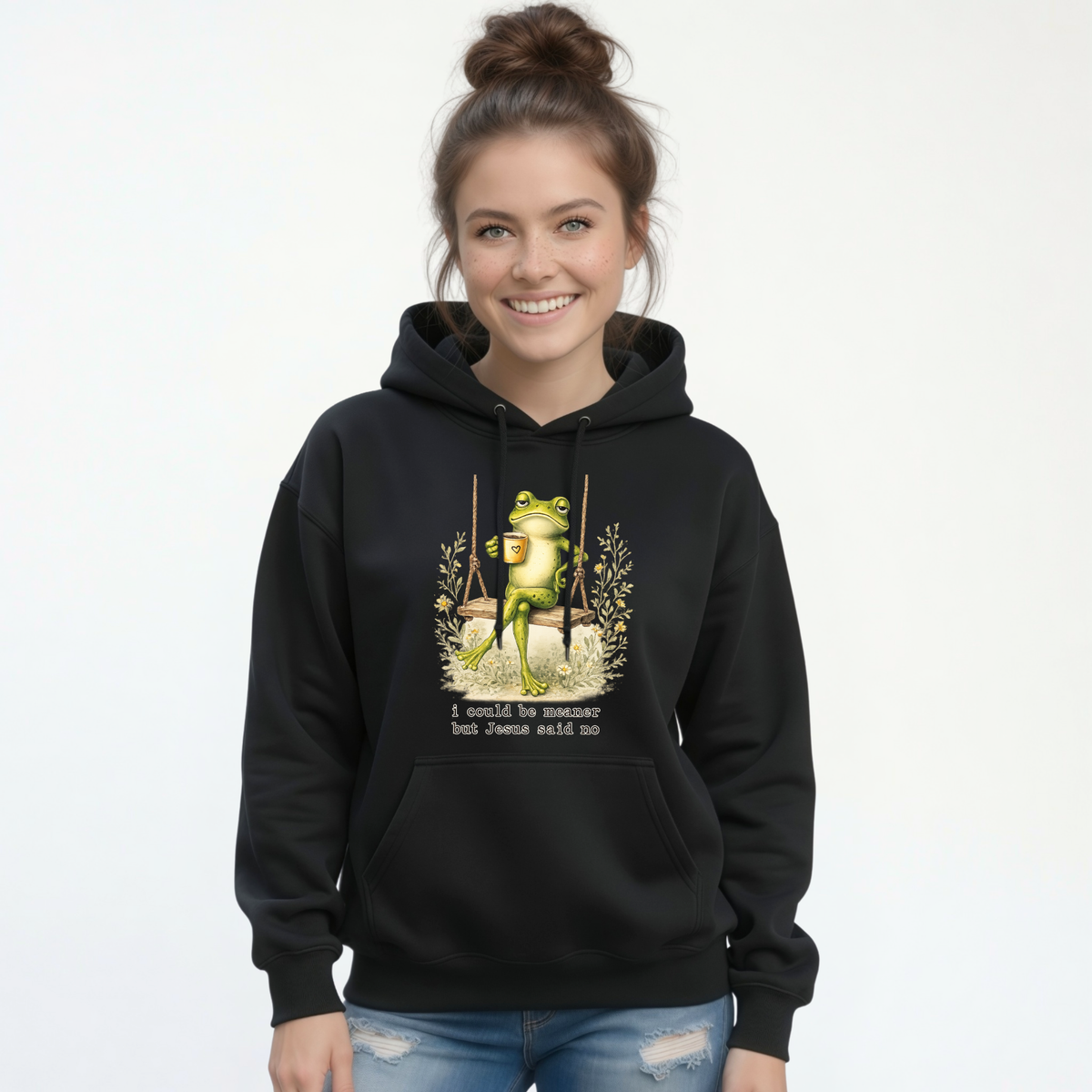 Woman modeling a black hoodie with a frog sitting on a swing holding a mug and the words “I could be meaner but Jesus said no.”