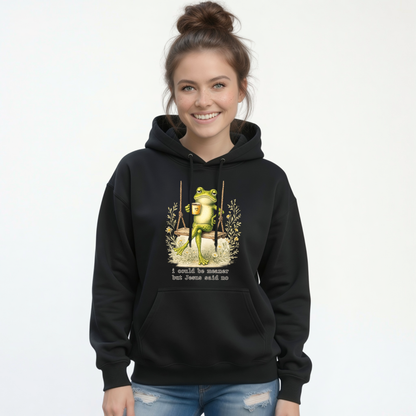Woman modeling a black hoodie with a frog sitting on a swing holding a mug and the words “I could be meaner but Jesus said no.”