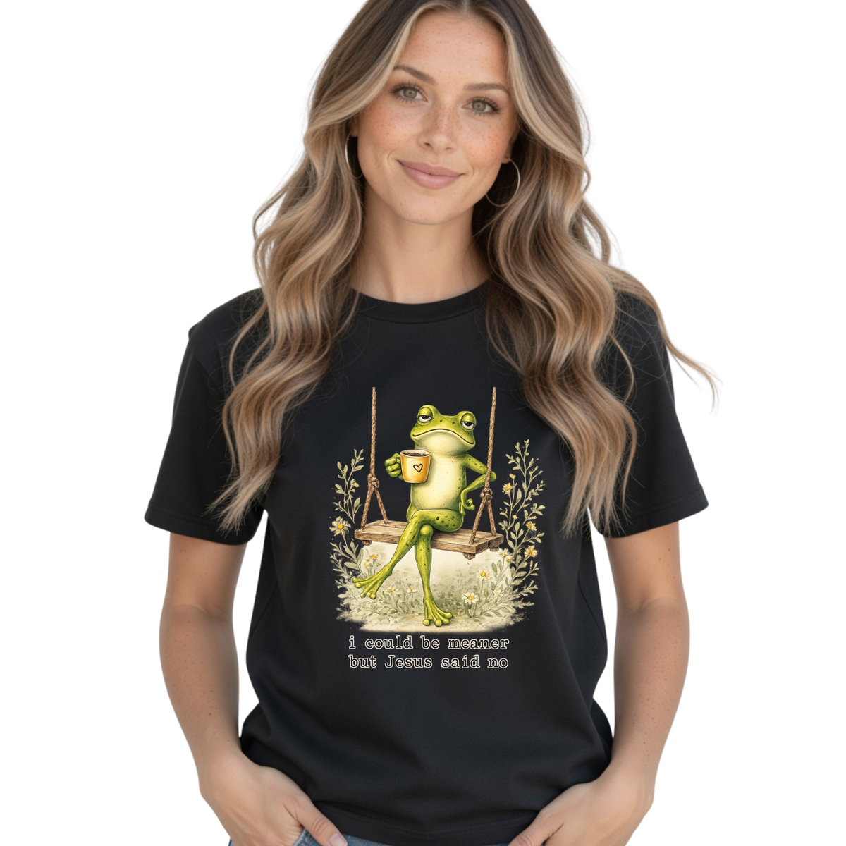 Woman modeling a black T-shirt with a frog sitting on a swing holding a mug and the words “I could be meaner but Jesus said no.”